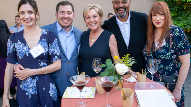 A group of five guests gather around a cocktail table outdoors, enjoying wine and conversation under the evening light at NBCC’s A Night in Tuscany benefit.