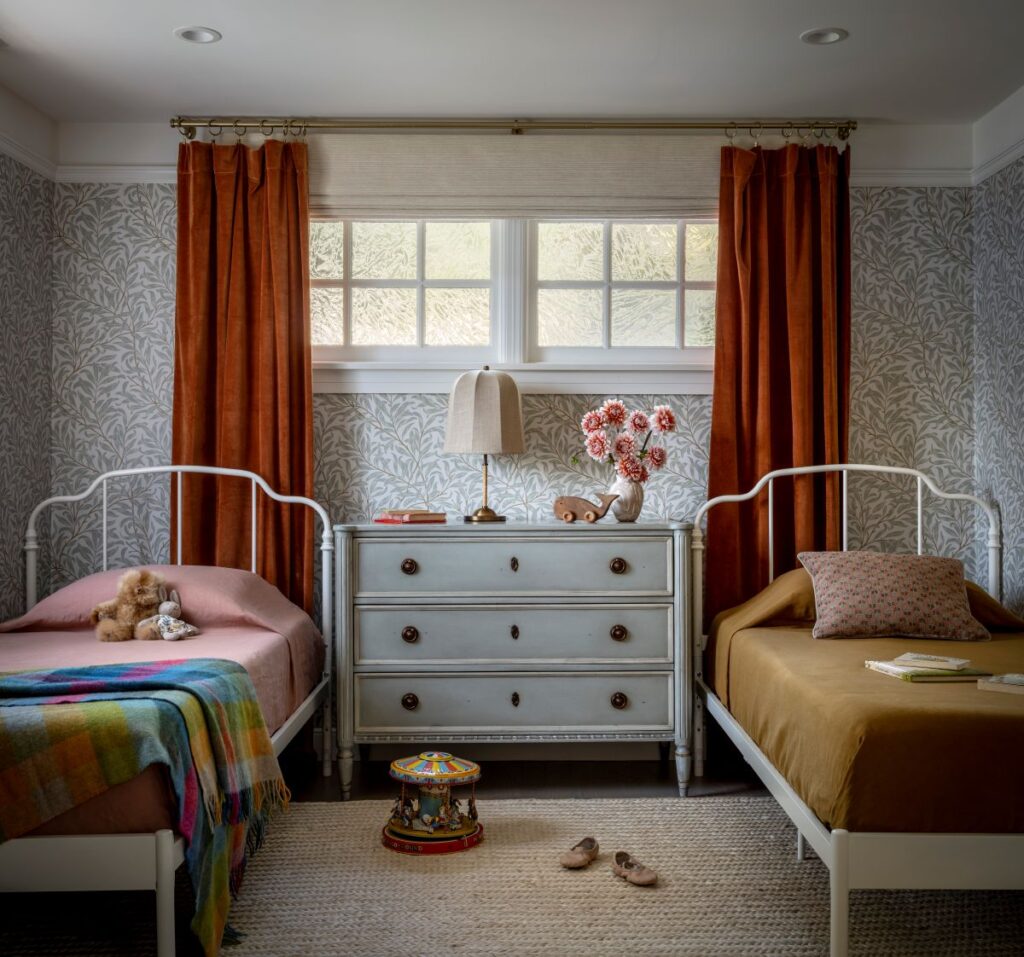 Twin girls’ bedroom with vintage-inspired white beds, botanical wallpaper, rust-colored velvet curtains, a shared pale blue dresser, and playful decorative accents.