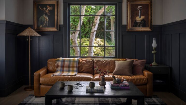 Cozy charcoal-paneled den featuring a caramel leather sofa, whimsical portraits of an ostrich and moose in Elizabethan attire, and a black coffee table with layered textures.