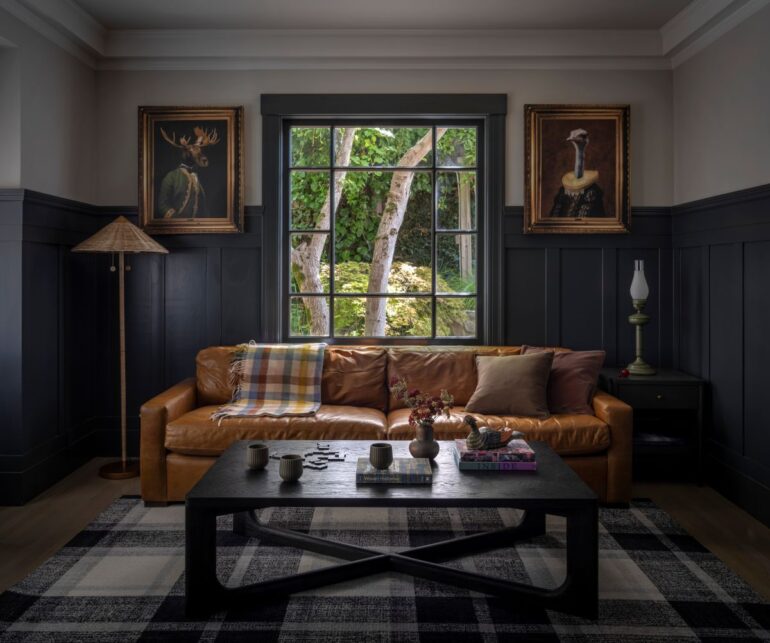 Cozy charcoal-paneled den featuring a caramel leather sofa, whimsical portraits of an ostrich and moose in Elizabethan attire, and a black coffee table with layered textures.