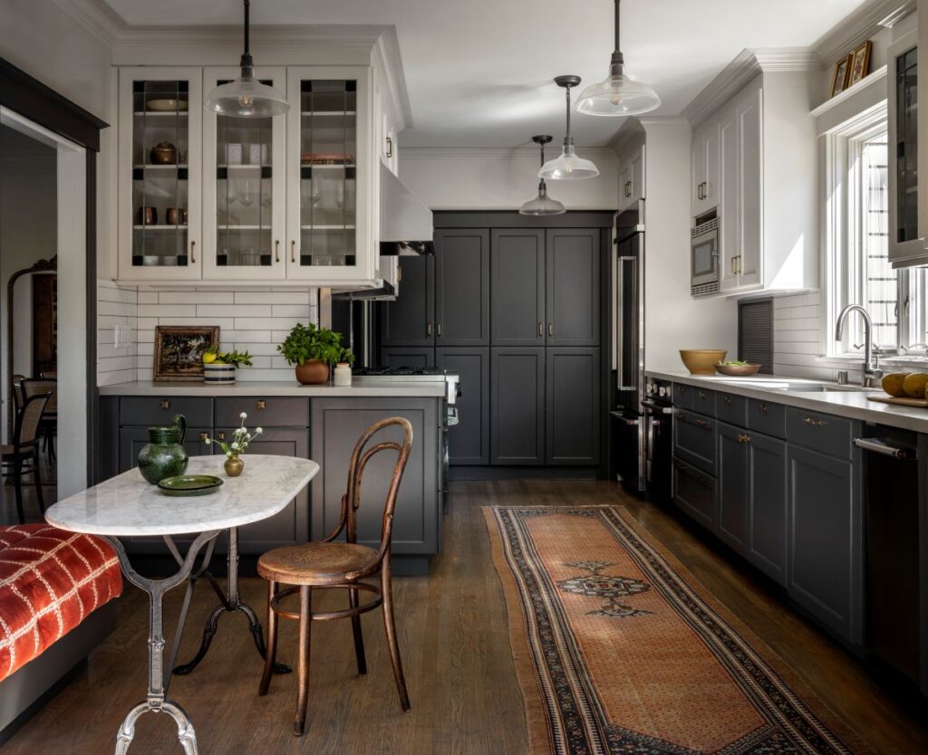 Renovated kitchen with ivory upper cabinets, charcoal lower cabinets, antique runner, marble breakfast nook with a red built-in bench, and pendant lighting overhead.