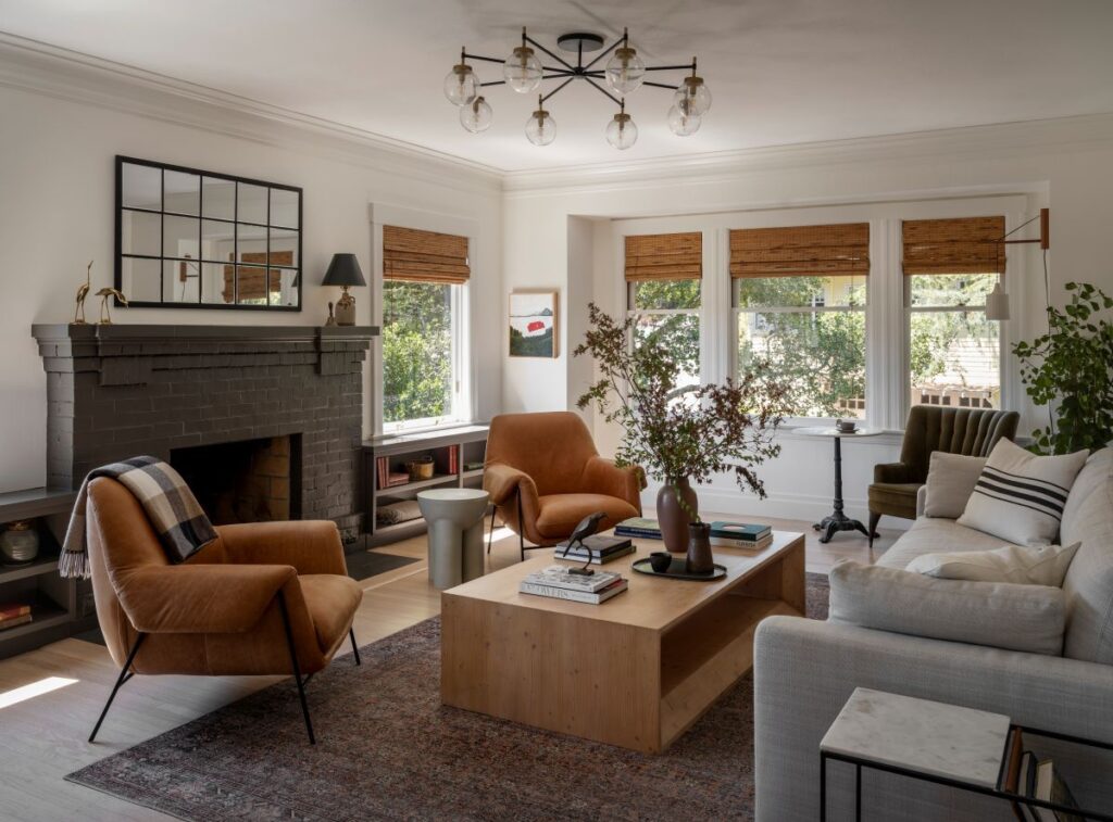 Living room blending modern and vintage pieces, with caramel leather armchairs, a neutral sofa, black brick fireplace, wood coffee table, and large windows with woven blinds.