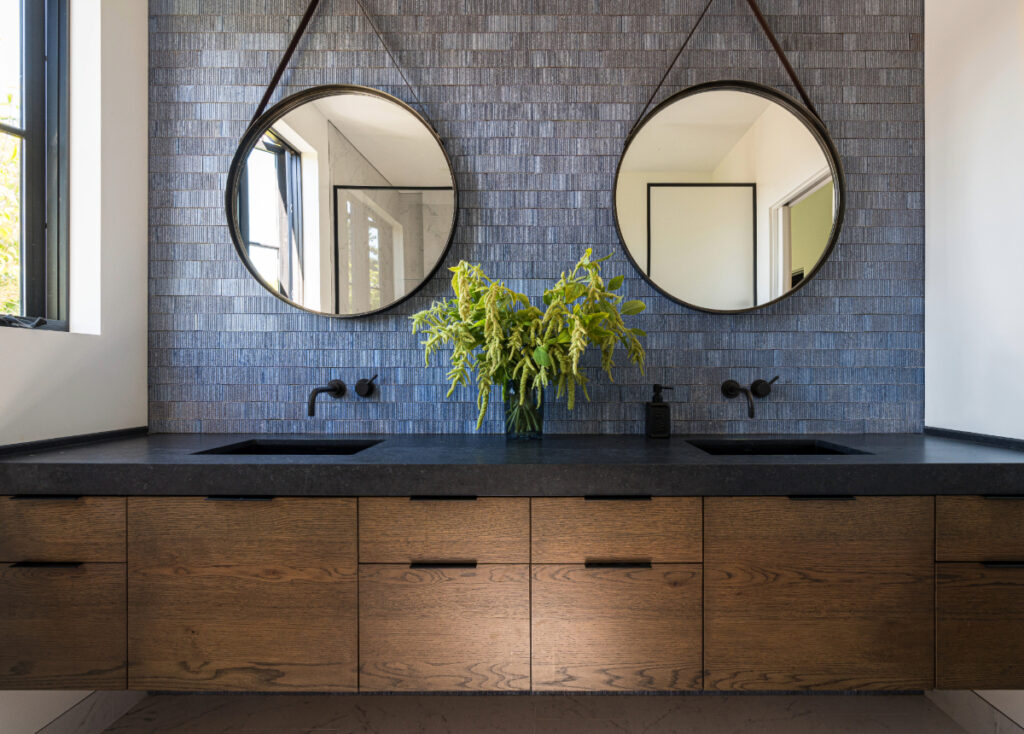 Primary bathroom with a floating oak vanity, black countertop, dual round mirrors, and handmade blue textured tile backsplash, accented by natural light from side windows.