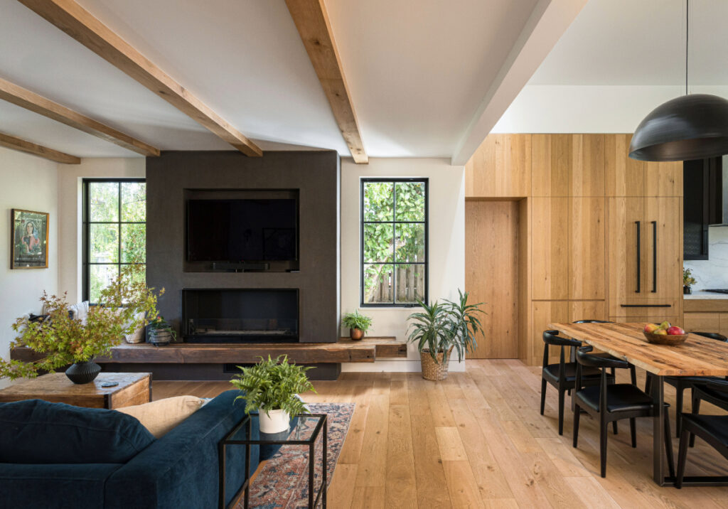Living room with exposed wood ceiling beams, a dark gray plaster fireplace wall with a mounted TV, recycled wood hearth, and large windows bringing in natural light, adjacent to a dining area with custom wood cabinetry.