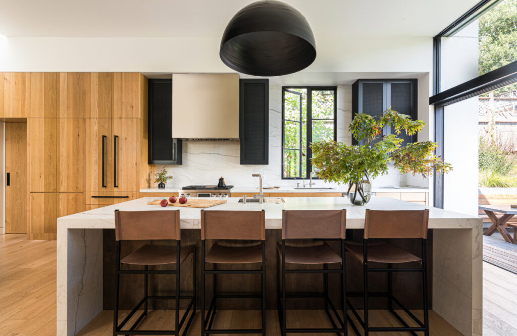 Modern kitchen featuring a large marble waterfall island with seating for four, custom white oak cabinetry paired with black upper cabinets, and expansive windows connecting the space to the outdoor dining area.