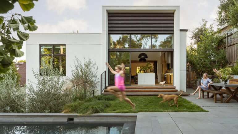 Backyard view of the renovated San Anselmo home with sliding glass doors opening to the kitchen, a child running with a dog on the grass, a woman seated at a wooden picnic table, and a pool in the foreground.