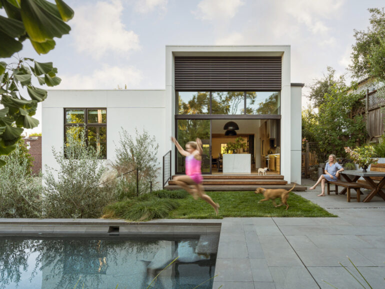 Backyard view of the renovated San Anselmo home with sliding glass doors opening to the kitchen, a child running with a dog on the grass, a woman seated at a wooden picnic table, and a pool in the foreground.