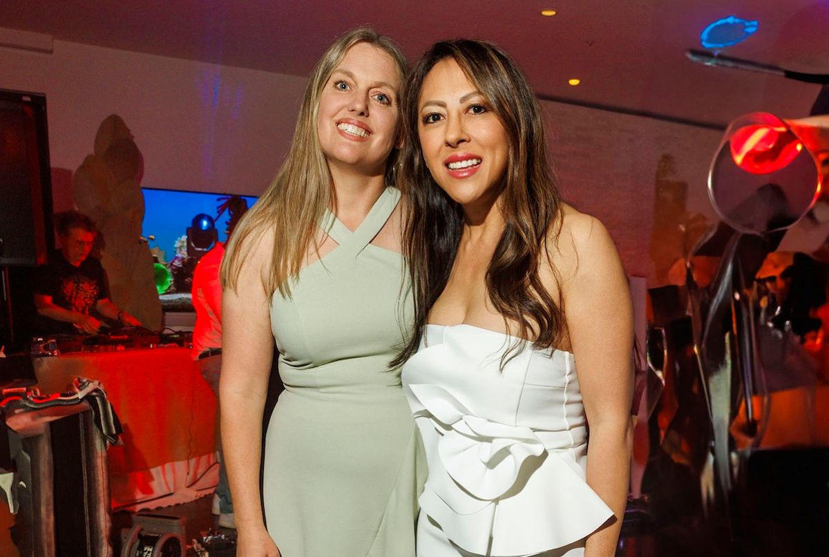 Two women smiling together at the event, one in a light green dress and the other in a strapless white dress, with a DJ booth in the background.