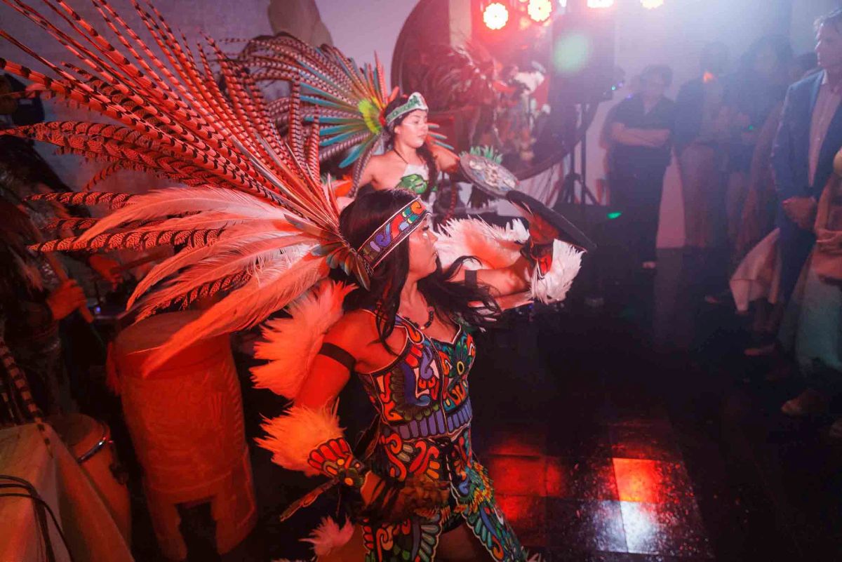 Performers in vibrant feathered headdresses and traditional Aztec attire dance under red stage lighting during the Belvedere-Hawthorne Nursery Schools fundraiser.