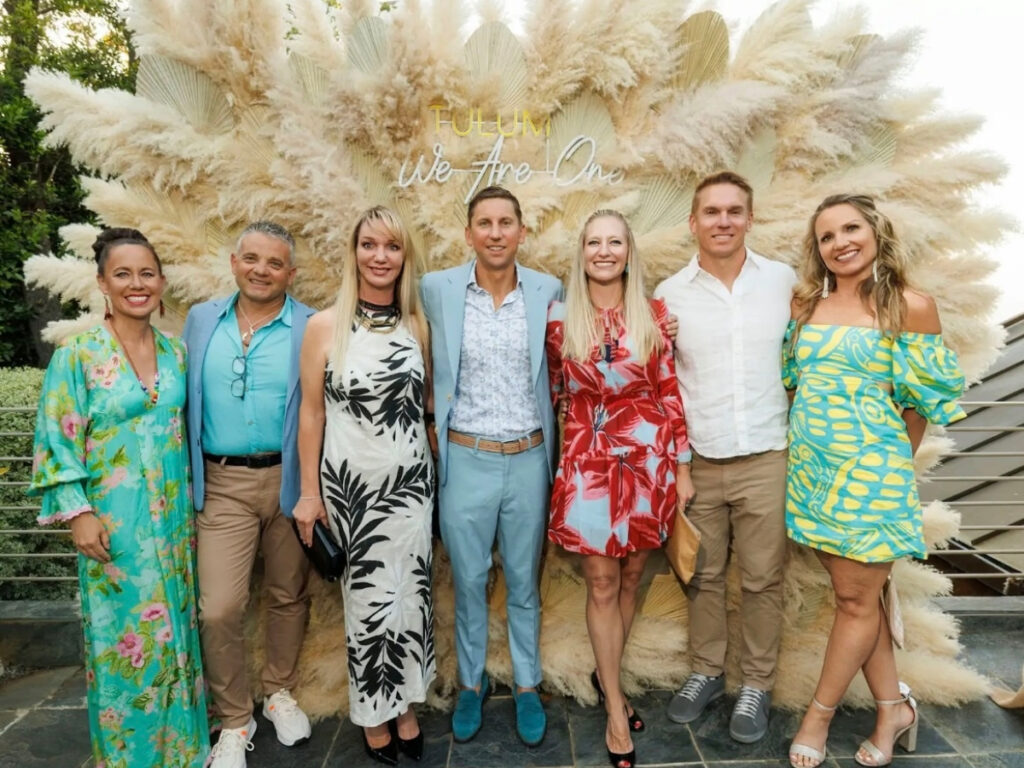 Seven guests pose in colorful attire in front of a large pampas grass installation with a neon sign reading “Tulum: We Are One.”