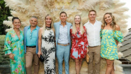 Seven guests pose in colorful attire in front of a large pampas grass installation with a neon sign reading “Tulum: We Are One.”