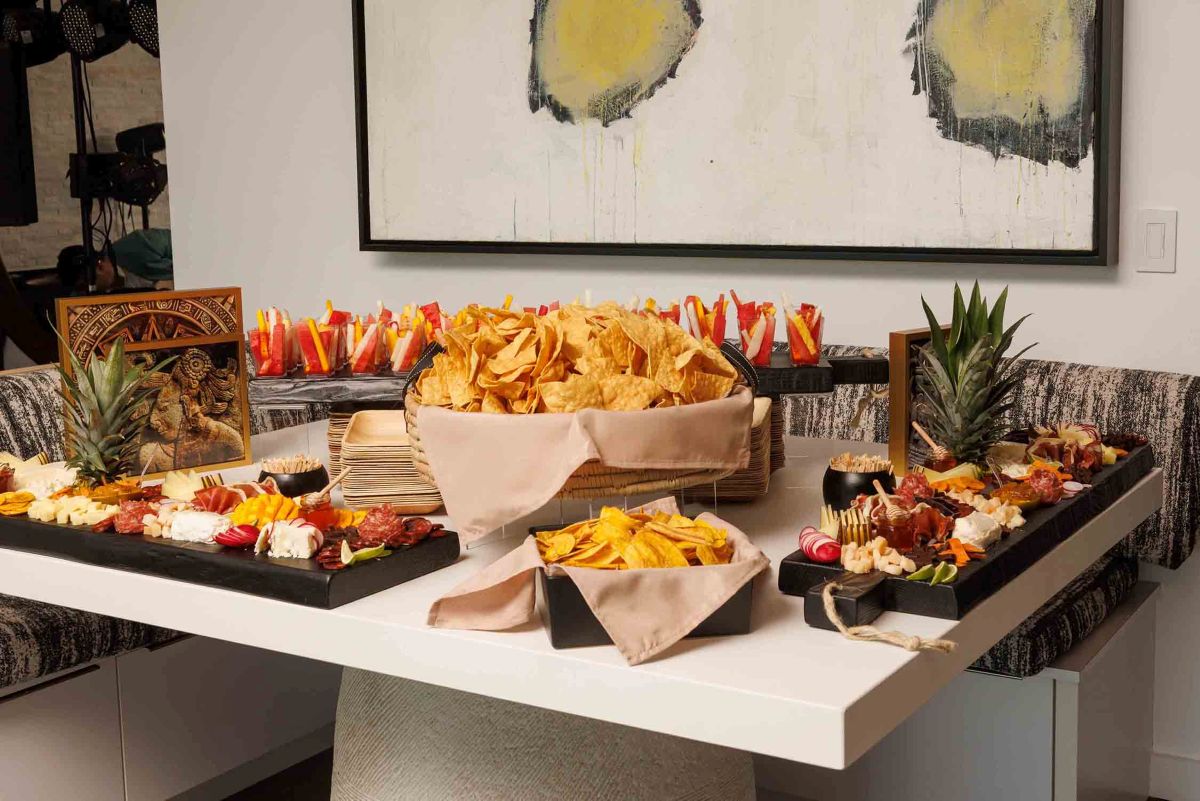 A buffet table with baskets of tortilla chips, fresh fruit, cheese, and charcuterie, accented with pineapples and decorative art pieces.