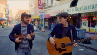 Two musicians strumming guitars on a lively city street, attracting the attention of nearby pedestrians.
