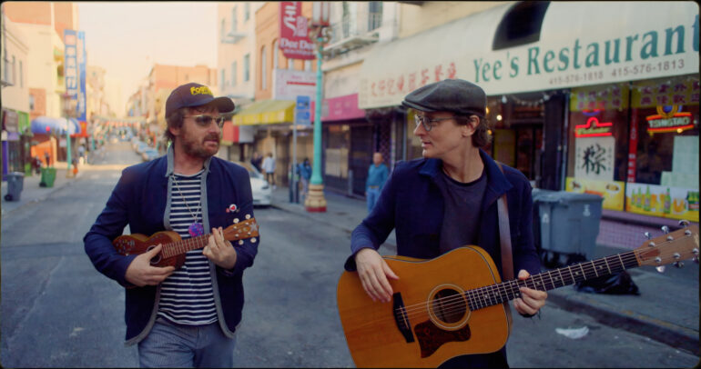 Two musicians strumming guitars on a lively city street, attracting the attention of nearby pedestrians.