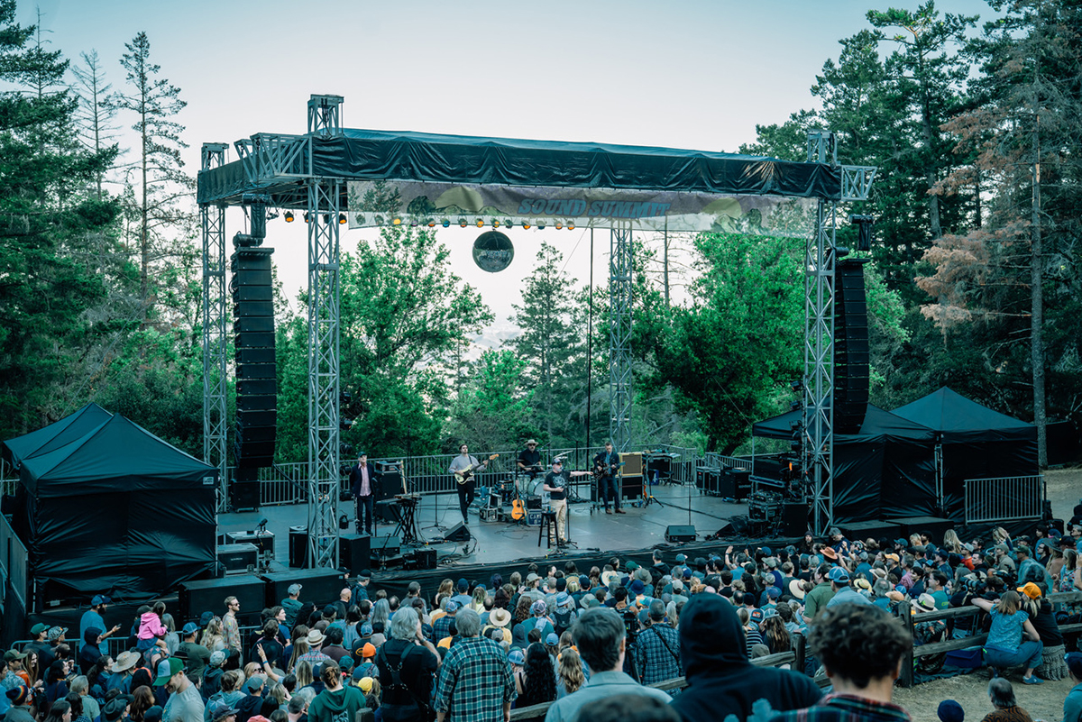 A vibrant crowd gathered at a concert in the woods, with people enjoying the music amidst lush greenery.