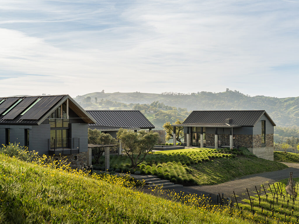 A house perched on a hill, overlooking a lush vineyard in the background under a clear blue sky.