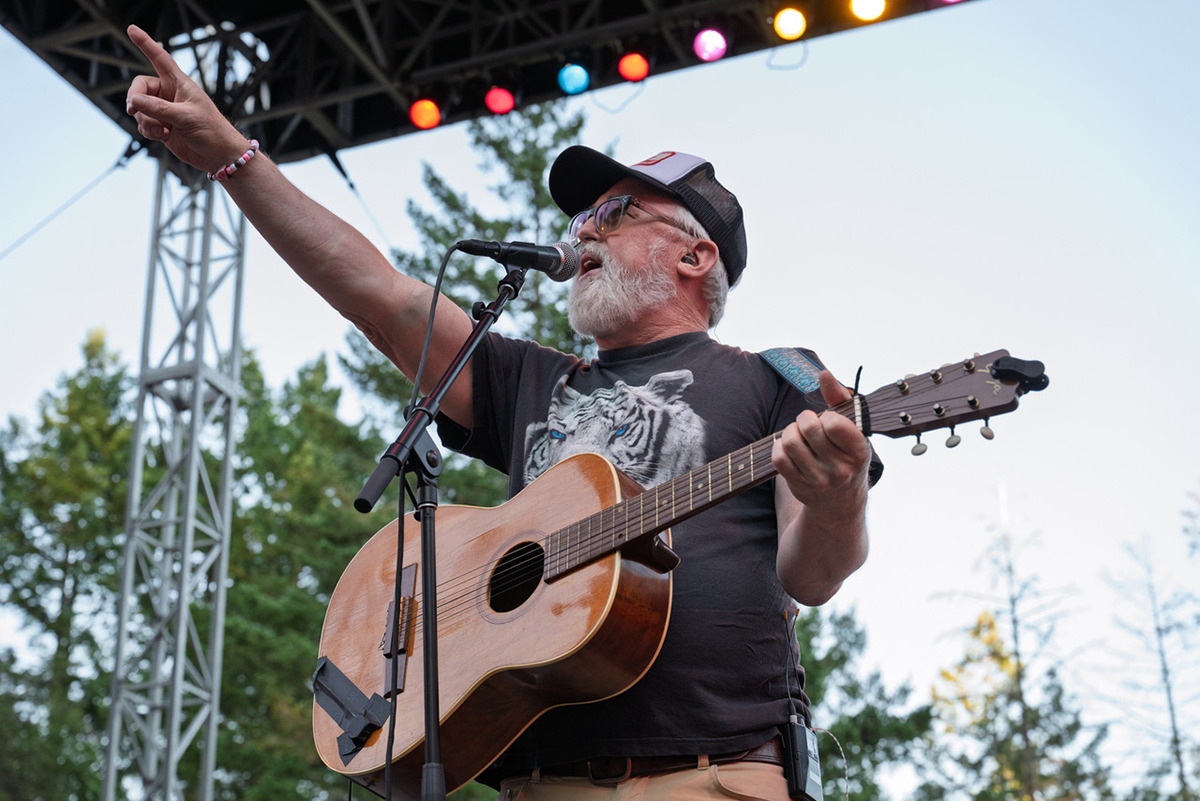 A bearded man wearing a hat, John McCrea of CAKE, plays an acoustic guitar during a live performance.