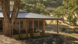 A house with a metal roof perched on a grassy hill, surrounded by trees and a clear blue sky.