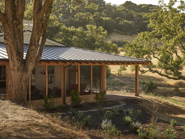 A house with a metal roof perched on a grassy hill, surrounded by trees and a clear blue sky.