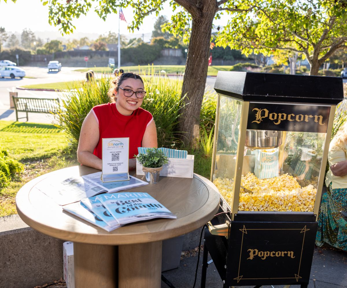 Smiling woman at popcorn booth with vintage machine and magazines.