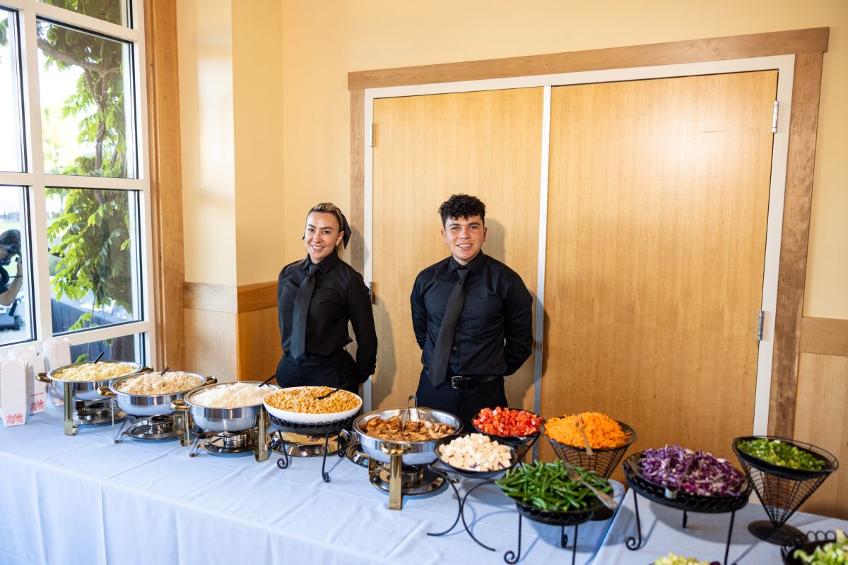Servers smiling behind buffet table with assorted dishes and toppings.