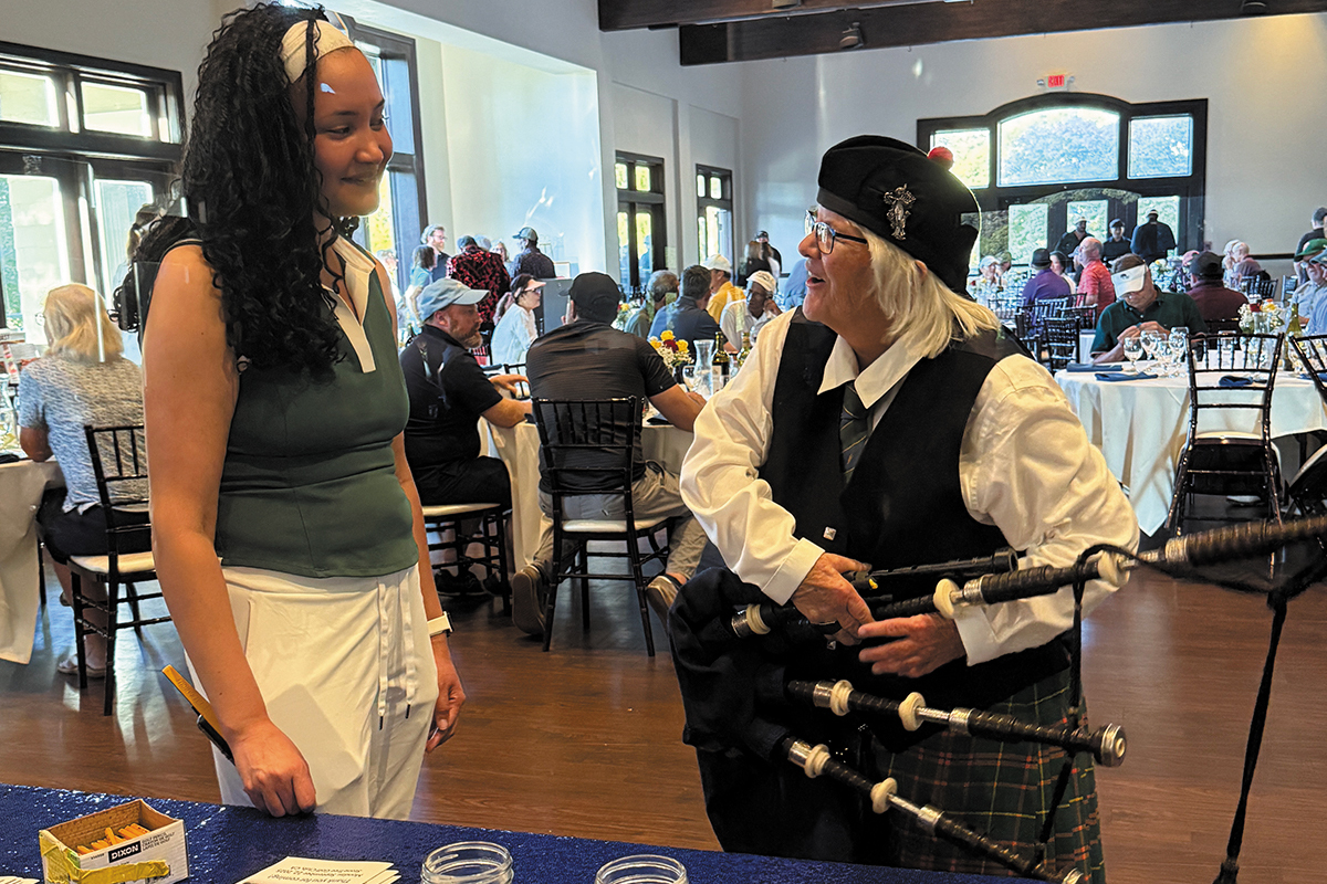 Two women talk at an indoor event; one holds bagpipes and wears traditional Scottish attire.