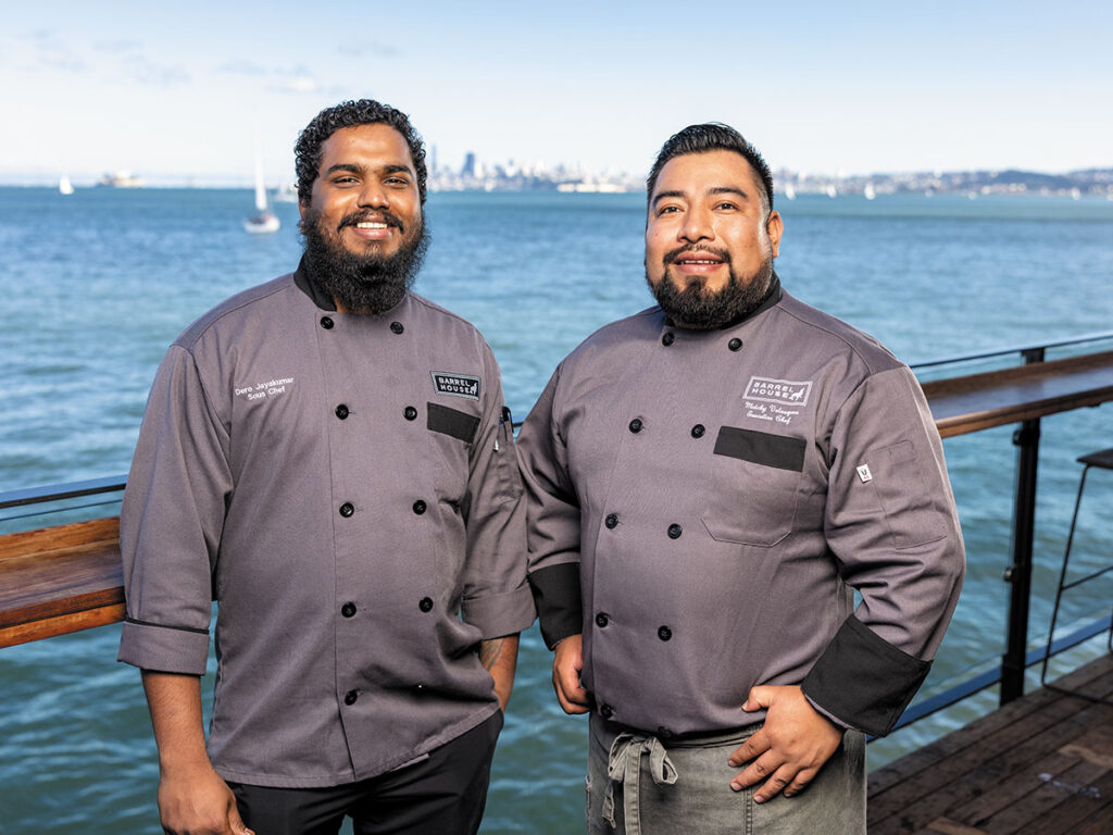 Two chefs stand side by side on a dock, wearing uniforms and smiling at the camera.