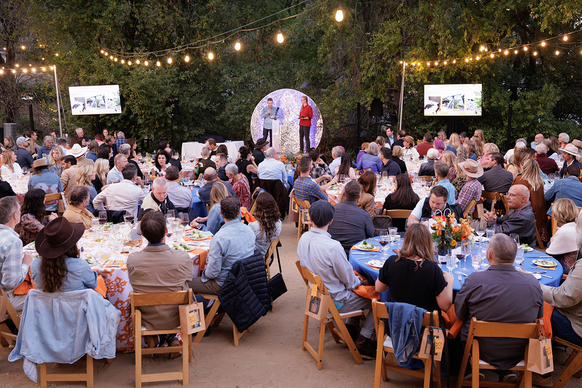 A large group of people seated at tables in a vibrant outdoor setting, enjoying food and conversation.