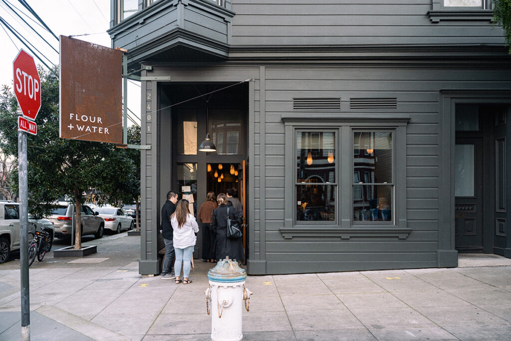 Two people are standing outside a restaurant, smiling and chatting, with outdoor seating visible nearby.