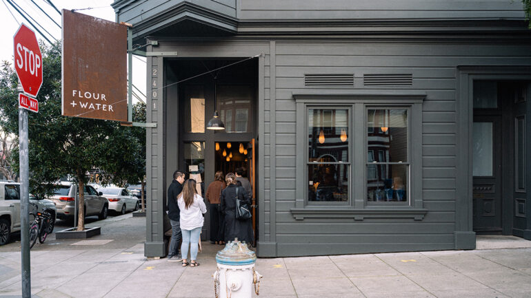 Two people are standing outside a restaurant, smiling and chatting, with outdoor seating visible nearby.