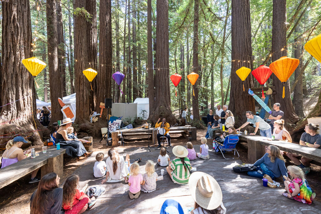 Children sitting at tables among trees in the woods, participating in a social event or meeting outdoors.