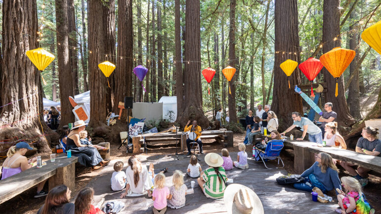 Children sitting at tables among trees in the woods, participating in a social event or meeting outdoors.