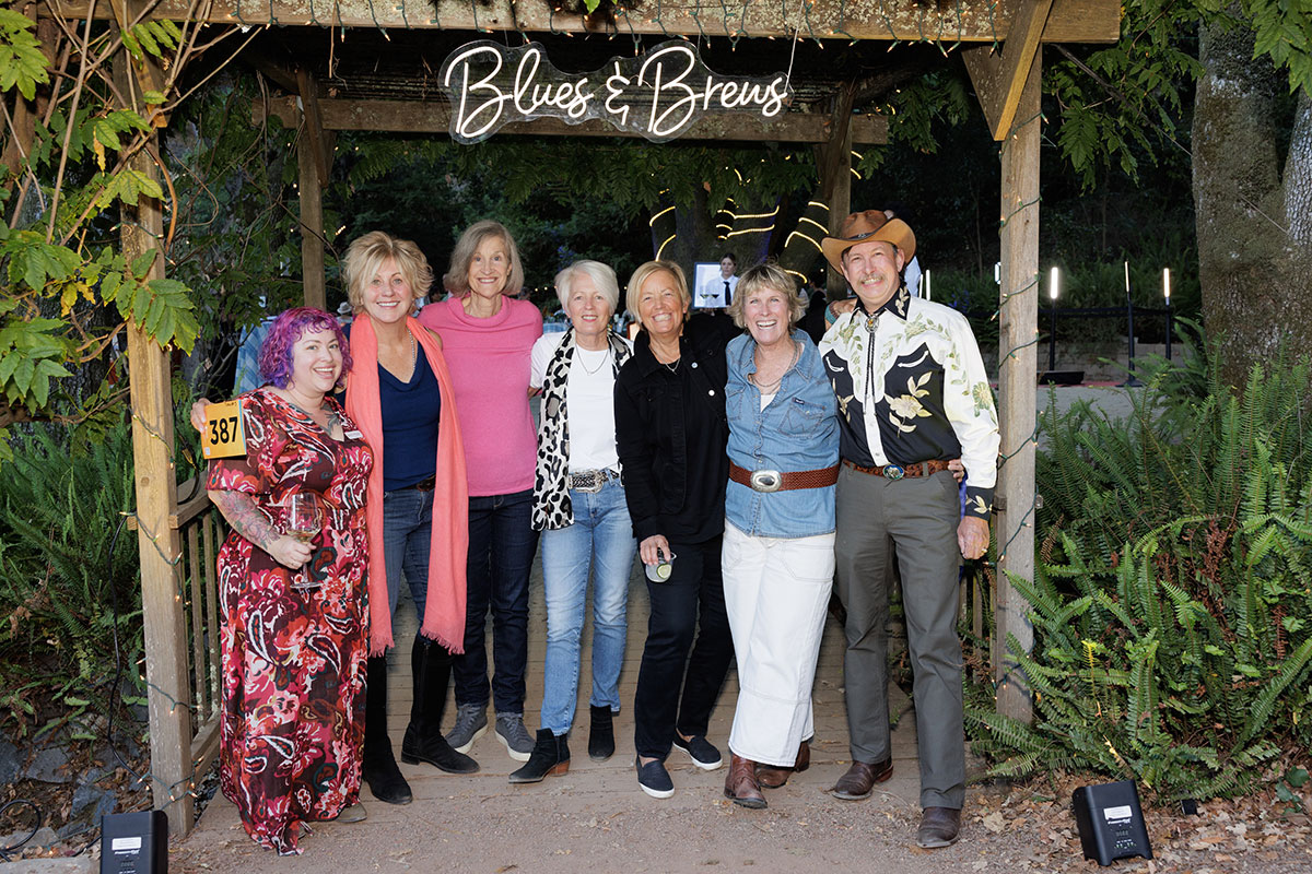 A group of people smiling and posing together in front of a large sign "Blues & Brews" , capturing a moment of camaraderie.