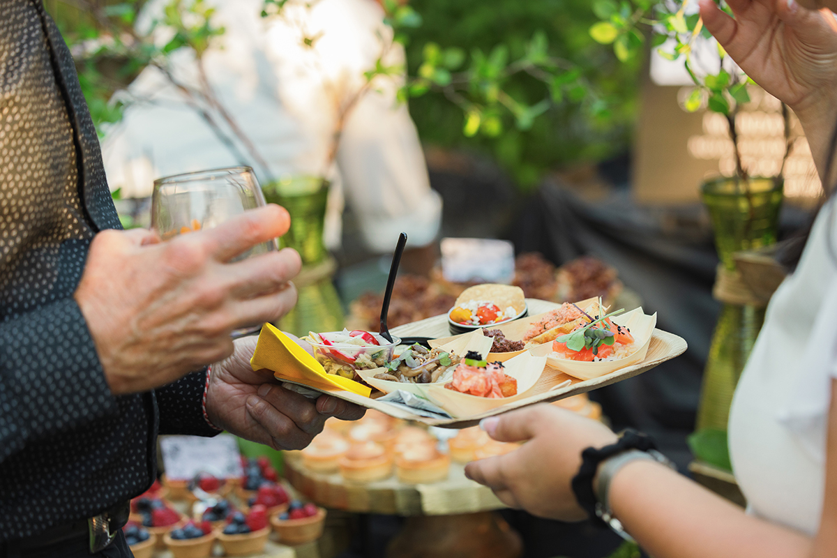 A man and woman stand together, each holding a tray of food, ready to serve at a social event.