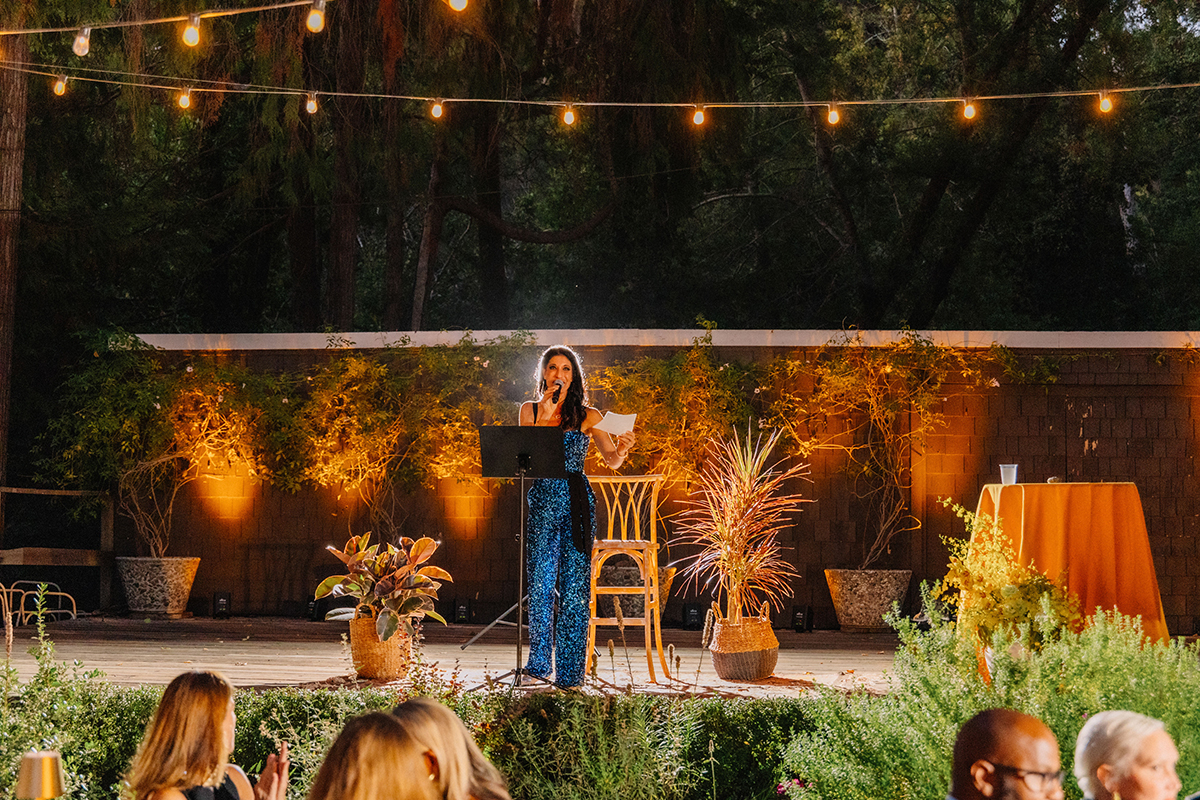 A woman delivering a speech at an outdoor event, surrounded by guests and decorated with flowers and greenery.