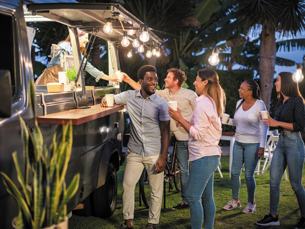 A diverse group of people gathered around a food truck, engaging in conversation and enjoying the outdoor dining experience.