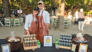 A couple stands beside a table displaying several bottles of wine, smiling and enjoying each other's company.