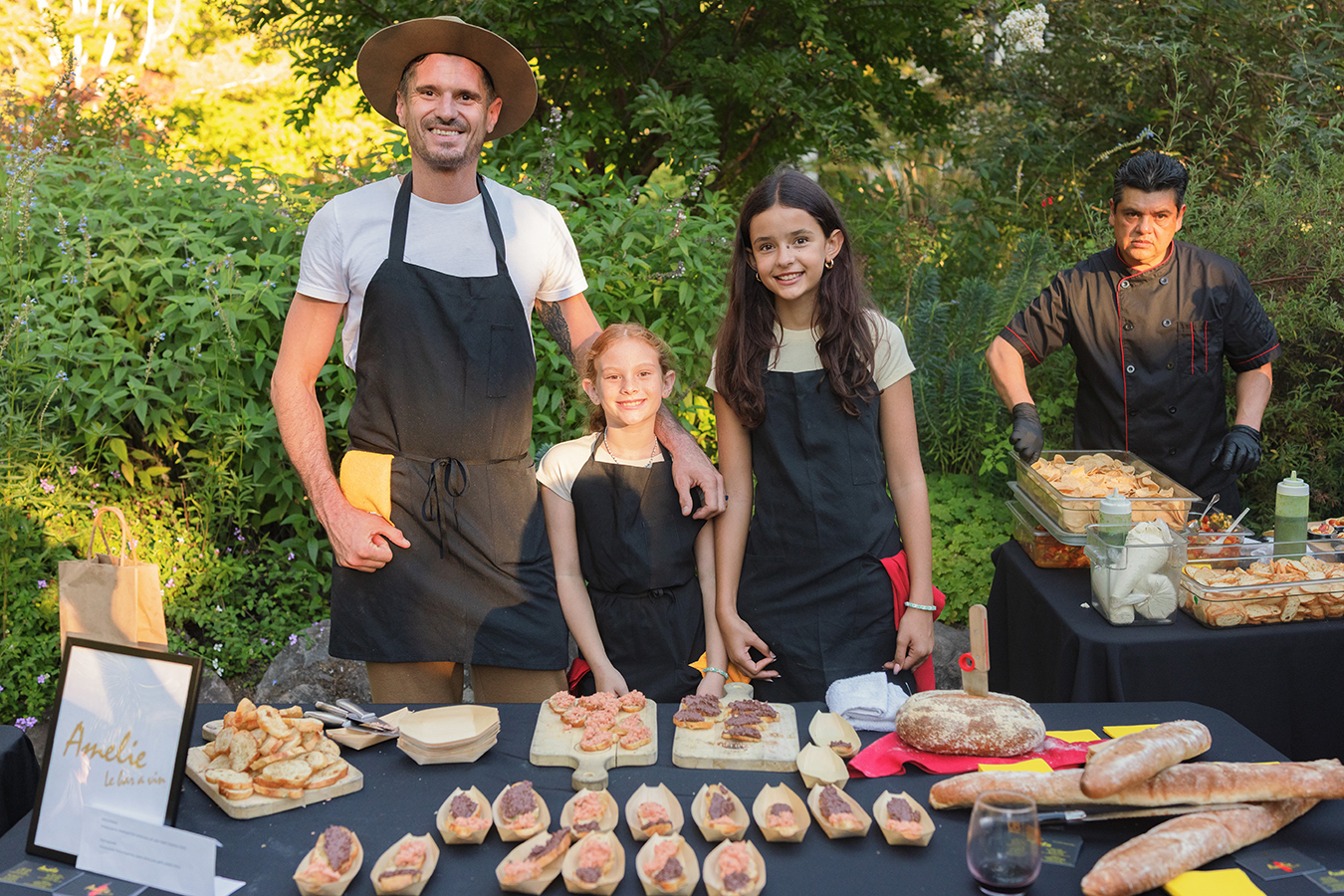 A man and two young girls in aprons stand behind a food stall with bread and appetizers at an outdoor event.
