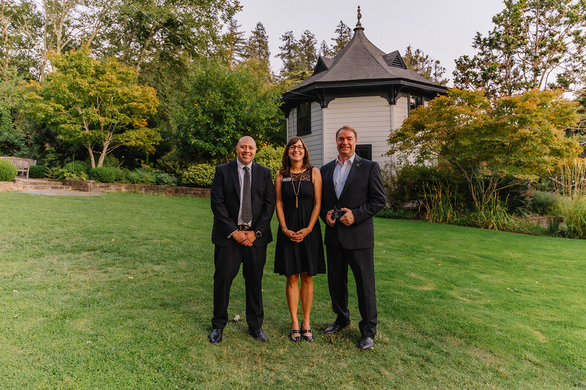 Three individuals dressed in suits pose in front of a residential house, suggesting a business meeting or event.