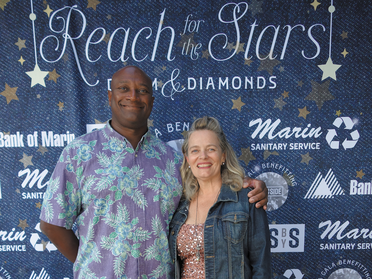 A man and woman pose together in front of a "Reach for the Stars" banner, smiling and looking optimistic.