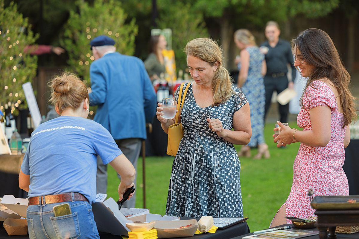 Two women, one wearing a blue dress and the other a red dress stand beside a table filled with various dishes and food items.