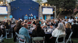 A large crowd of people seated at tables enjoying a gathering in a grassy field under a clear sky.