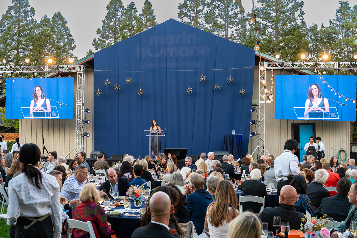 A woman speaking at an outdoor event, gesturing passionately while addressing a crowd gathered around her.
