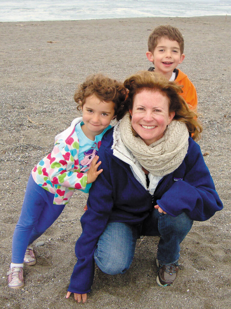 A woman stands with two children on a beach, surrounded by sand and waves, sharing a joyful moment.