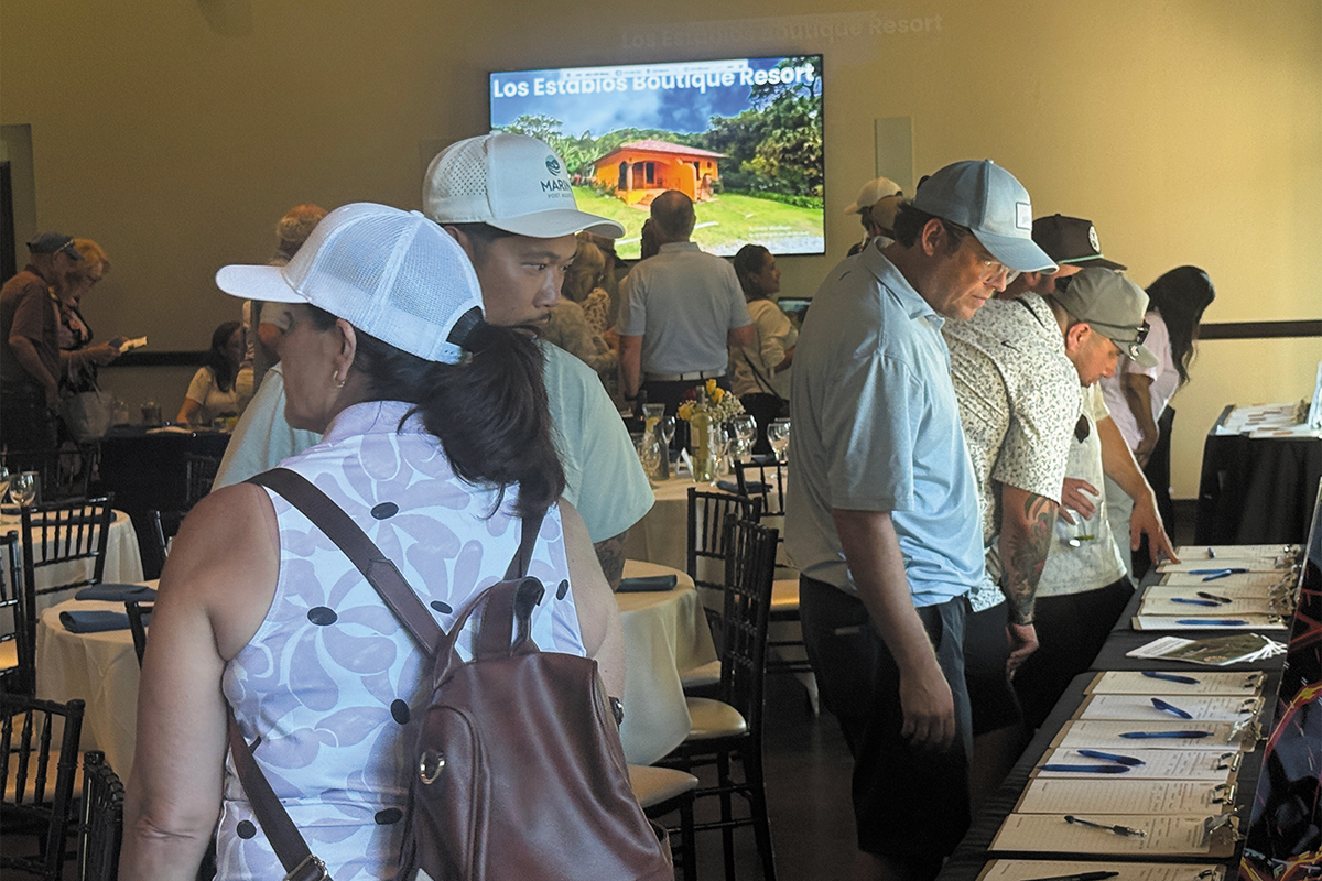 A group of people wearing hats and shirts gathered around a table full of papers with a television in the background.