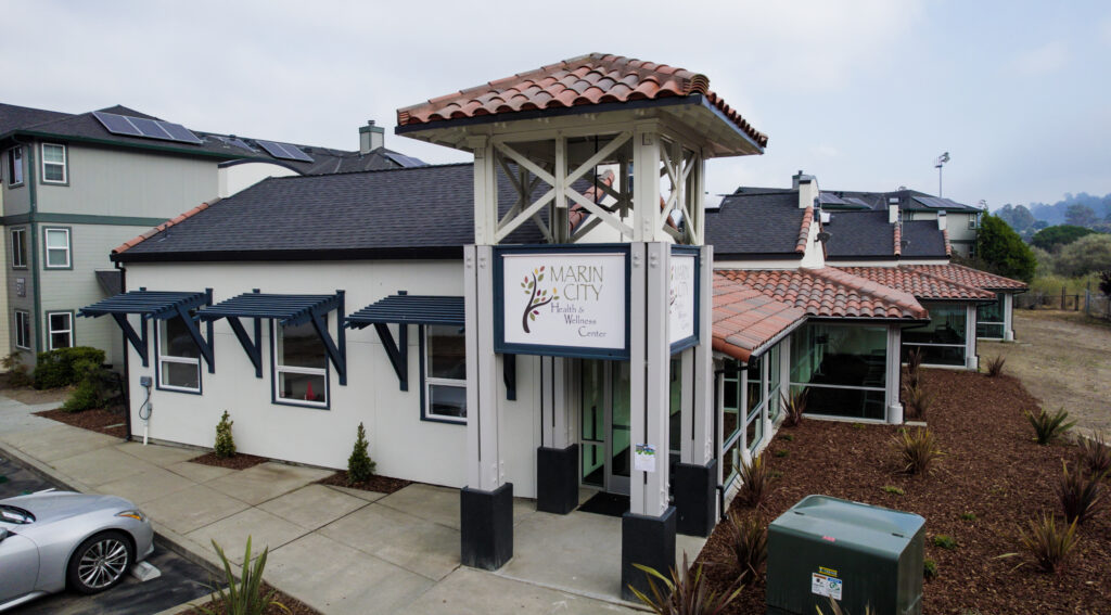 The exterior of Marin City Heath and Wellness center is featured. It is a white building with a black roof and a two story tower that houses the sign.