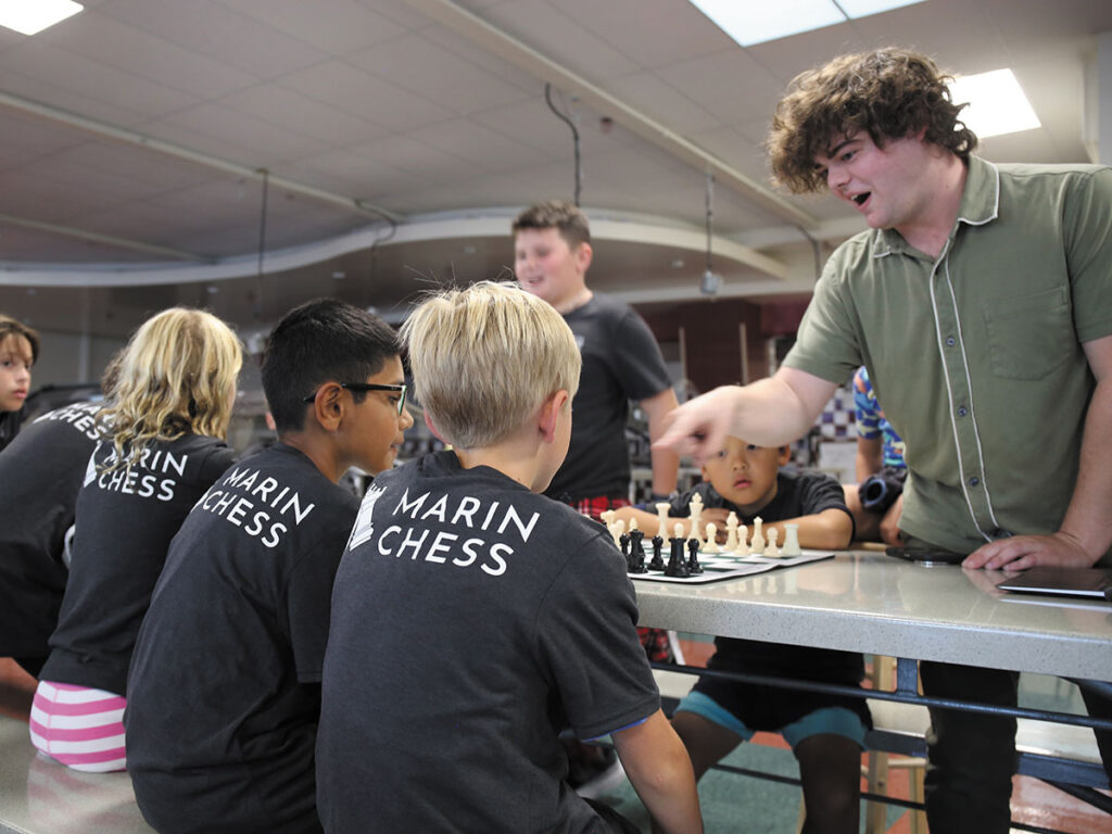A man instructs a group of children on how to play chess, demonstrating strategies and moves on a chessboard.