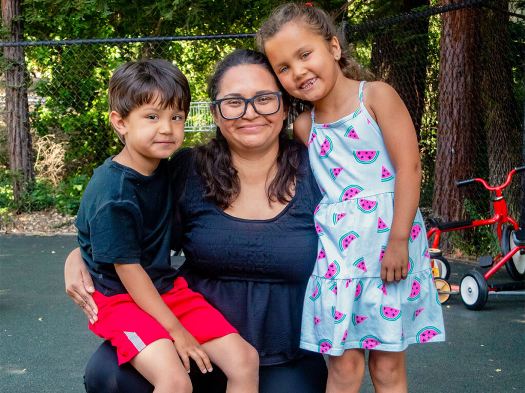 A woman poses with two children on a playground and a sunny day in the background.