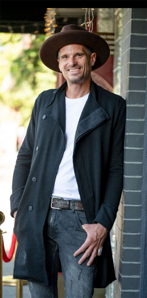 A man wearing a hat and coat stands outside, looking thoughtfully into the distance.