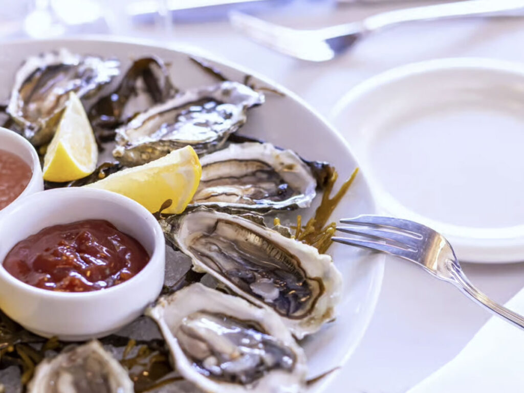 A platter of oysters on the half shell garnished with lemon slices and served with a small bowl of cocktail sauce.